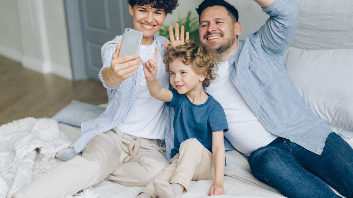 Happy family mom dad and child taking selfie in bed waving hands smiling